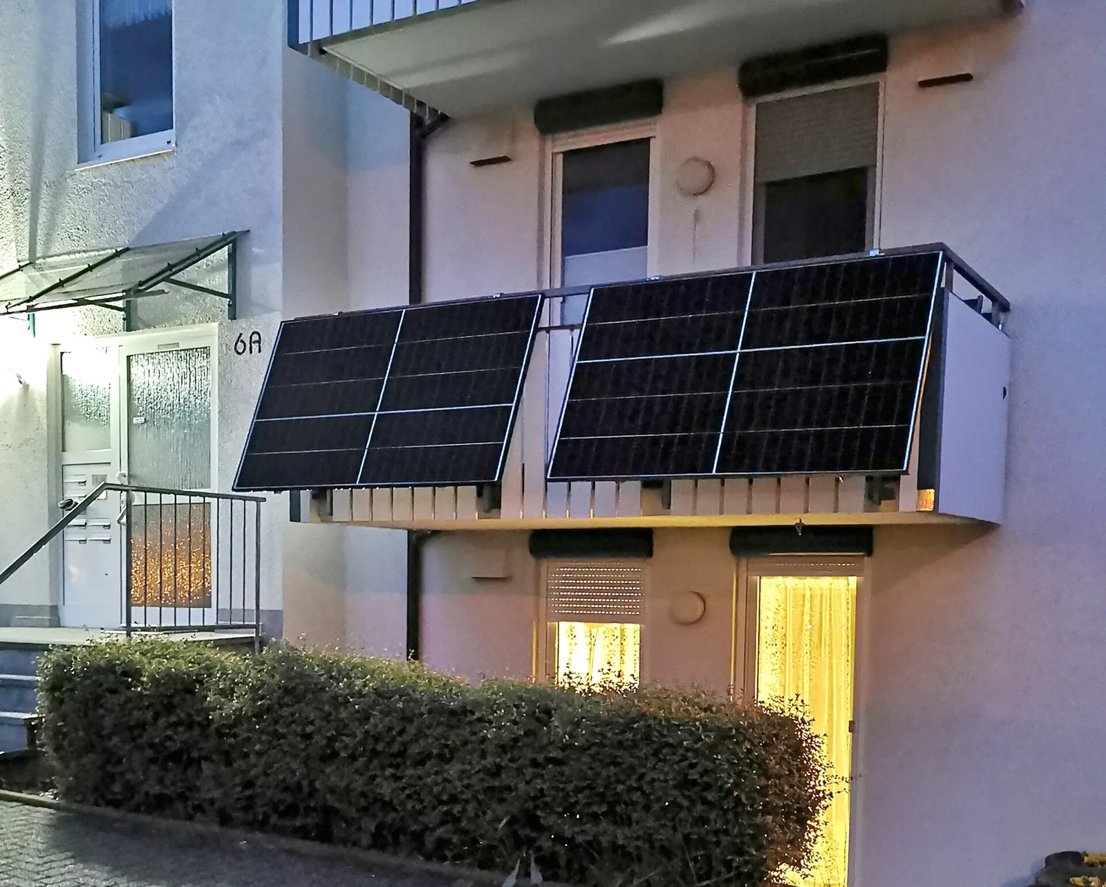 Balkon eines Wohnhauses in der Dämmerung mit zwei großen Solarmodulen an der Brüstung, davor eine Hecke.