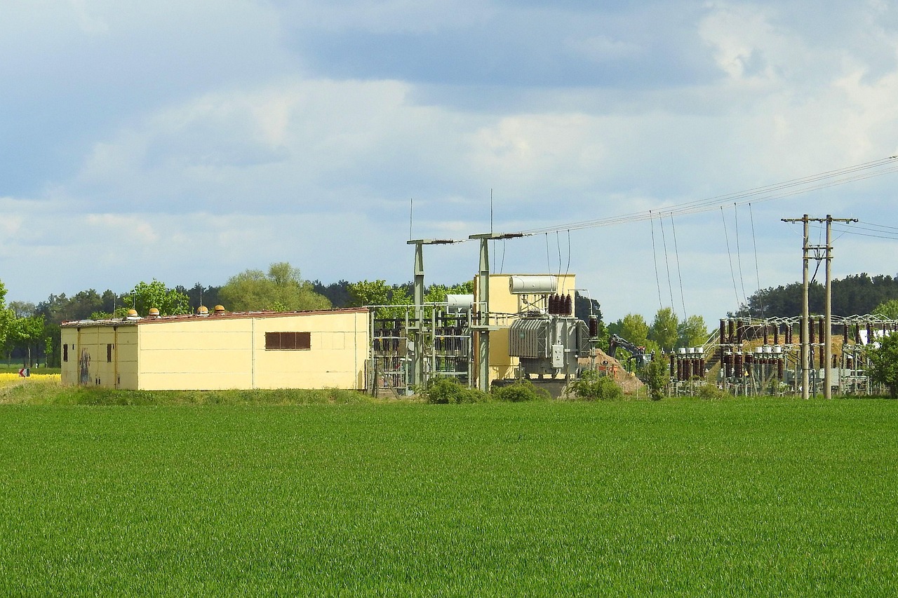 Umspannwerk mit Transformator und Freileitungsmasten am Rand eines Getreidefeldes unter blauem Himmel.