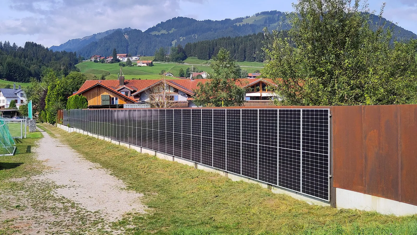 Senkrecht montierte Solarmodule als Zaun an einem Grundstück mit Blick auf Häuser und Berge.