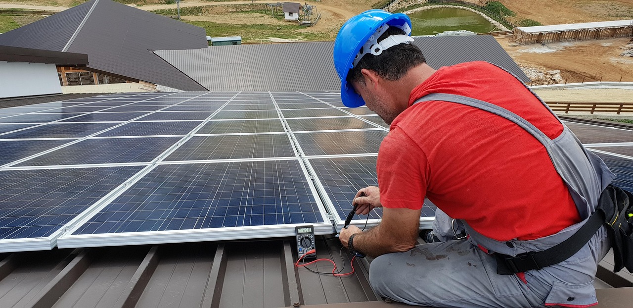 Techniker in rotem Shirt und blauem Schutzhelm kniet auf einem Blechdach und misst mit einem Multimeter die elektrischen Werte eines Photovoltaikmoduls
